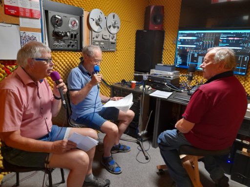 Drei Männer im Studio sprechen ins Mikrofon und verwenden Notizen. Technik im Hintergrund.